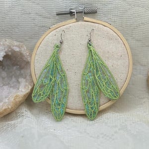 Può includere: Un paio di orecchini a forma di ali di fata verdi e iridescenti. Gli orecchini sono realizzati in un materiale delicato e merlettato e presentano dettagli intricati. Sono appesi a ganci color argento e sono esposti su un tessuto dai toni neutri all'interno di un telaio da ricamo.