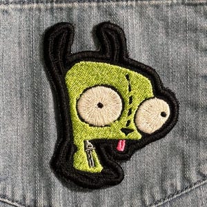 Puede incluir: Parche bordado con un personaje alienígena verde con grandes ojos blancos, una lengua rosa y un contorno negro. El parche está cosido sobre tela vaquera, mostrando un diseño de dibujos animados.