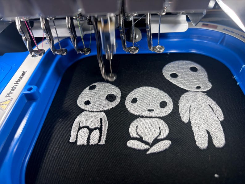 May include: An embroidery machine hoop holding black fabric with three silver-stitched figures. The figures are stylized, rounded, and feature simple facial expressions. The machine needles are positioned above the fabric, ready for stitching.