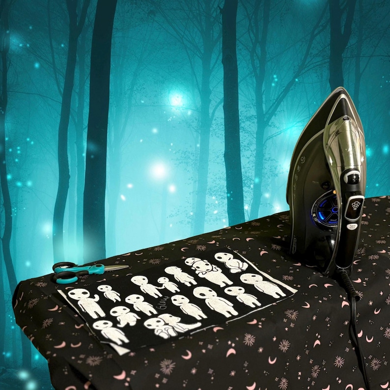May include: A black fabric sheet featuring a repeating white graphic print of small, stylized forest spirits, positioned on an ironing board with a black and silver electric iron and a pair of teal and black scissors resting nearby.
