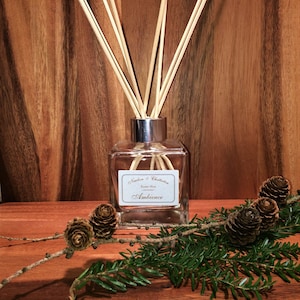 May include: A clear glass diffuser bottle with a silver cap and wooden sticks. The bottle has a label that reads "Nealson & Chattelton Essential Oils Ambience". The bottle is sitting on a wooden surface with pine branches and pine cones.