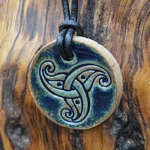 May include: A circular ceramic pendant featuring a teal and tan Celtic knot design, strung on a black leather cord. The pendant is about 4 cm in diameter. The backdrop is a textured wooden surface.