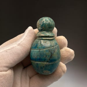 Könnte beinhalten: Eine kleine, tealfarbene, geschnitzte Stein-Flasche mit abgerundetem Oberteil und leicht ausgestelltem Hals. Die Flasche hat eine glatte, polierte Oberfläche mit einigen sichtbaren Linien.