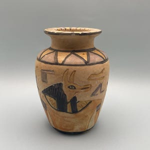 Puede incluir: Un pequeño jarrón de cerámica marrón con un diseño de inspiración egipcia. El jarrón presenta una representación en negro y marrón de un animal con un hocico largo y orejas grandes, rodeado de patrones geométricos.