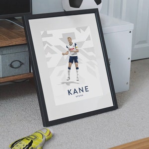 Pode incluir: Uma impress&atilde;o emoldurada de um jogador de futebol com um uniforme branco e azul com o texto "KANE SPURS" abaixo da imagem. O jogador est&aacute; em uma pose din&acirc;mica, com a perna direita levantada e o bra&ccedil;o esquerdo estendido. O fundo &eacute; um padr&atilde;o geom&eacute;trico branco e cinza.