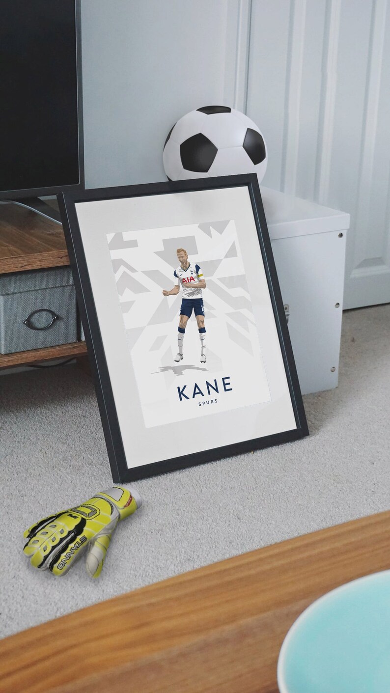 Pode incluir: Uma impress&atilde;o emoldurada de um jogador de futebol com um uniforme branco e azul com o texto "KANE SPURS" abaixo da imagem. O jogador est&aacute; em uma pose din&acirc;mica, com a perna direita levantada e o bra&ccedil;o esquerdo estendido. O fundo &eacute; um padr&atilde;o geom&eacute;trico branco e cinza.