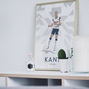 Pode incluir: Uma impress&atilde;o emoldurada de um jogador de futebol com uma camisa branca com detalhes azuis, celebrando um gol. O jogador est&aacute; vestindo shorts e meias. O fundo &eacute; um padr&atilde;o geom&eacute;trico branco e cinza. A impress&atilde;o &eacute; intitulada "Kane Spurs".