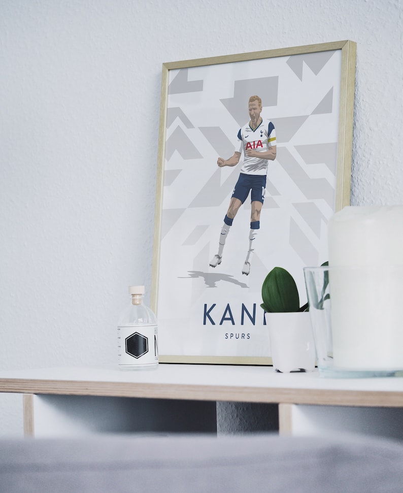 Pode incluir: Uma impress&atilde;o emoldurada de um jogador de futebol com uma camisa branca com detalhes azuis, celebrando um gol. O jogador est&aacute; vestindo shorts e meias. O fundo &eacute; um padr&atilde;o geom&eacute;trico branco e cinza. A impress&atilde;o &eacute; intitulada "Kane Spurs".
