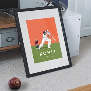 Pode incluir: Uma impress&atilde;o emoldurada de um jogador de cr&iacute;quete em a&ccedil;&atilde;o. O jogador est&aacute; vestindo um uniforme branco e um capacete. A impress&atilde;o tem um fundo vermelho e grama verde. O texto "KOHLI INDIA INTERNATIONAL CRICKET ICON" &eacute; impresso abaixo do jogador.
