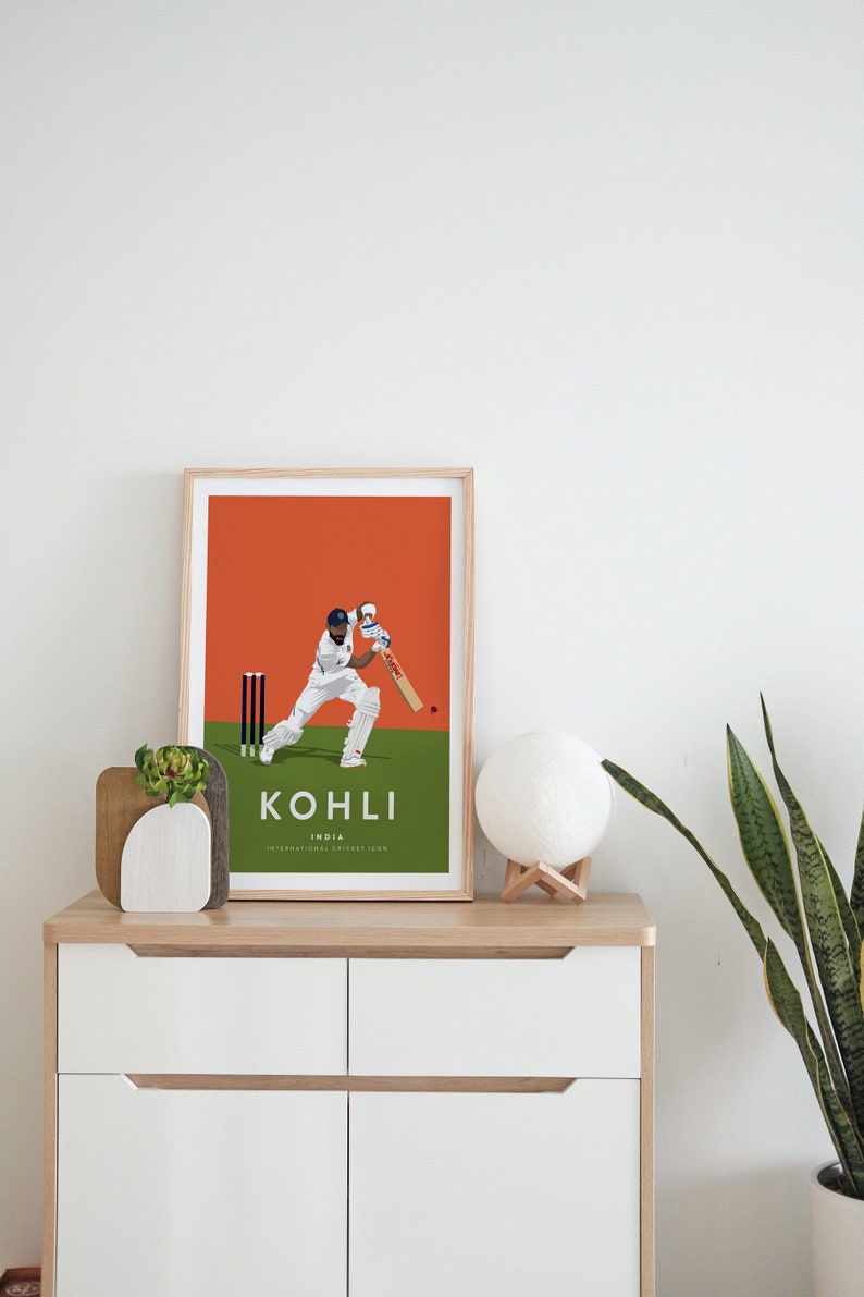 Pode incluir: Uma impress&atilde;o emoldurada de um jogador de cr&iacute;quete em a&ccedil;&atilde;o, com fundo laranja, grama verde e o texto "KOHLI INDIA INTERNATIONAL CRICKET ICON".