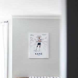 Pode incluir: Um p&ocirc;ster emoldurado branco com uma ilustra&ccedil;&atilde;o gr&aacute;fica de um jogador de futebol com um uniforme branco e azul marinho. O p&ocirc;ster tem o texto "KANE SPURS" na parte inferior.