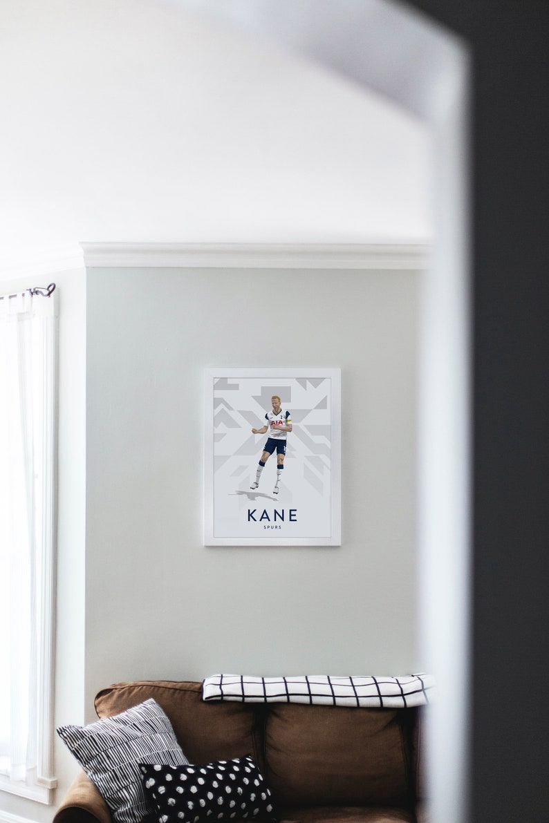 Pode incluir: Um p&ocirc;ster emoldurado branco com uma ilustra&ccedil;&atilde;o gr&aacute;fica de um jogador de futebol com um uniforme branco e azul marinho. O p&ocirc;ster tem o texto "KANE SPURS" na parte inferior.