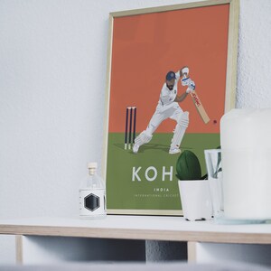 Pode incluir: Um p&ocirc;ster emoldurado com uma ilustra&ccedil;&atilde;o gr&aacute;fica de um jogador de cr&iacute;quete em a&ccedil;&atilde;o. O jogador est&aacute; vestindo um uniforme branco e um bon&eacute; azul. O p&ocirc;ster tem um fundo verde com um wicket de cr&iacute;quete e o texto "KOHLI INDIA INTERNATIONAL CRICKET".