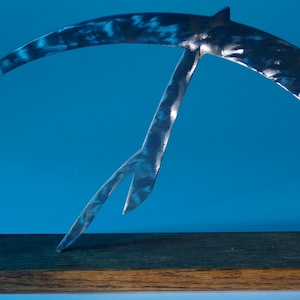 Op de afbeelding: Een zilveren metalen sculptuur met een gebogen vorm met een kleinere, puntige vorm die vanaf de bovenkant uitsteekt. Het beeld is gemonteerd op een houten voet.