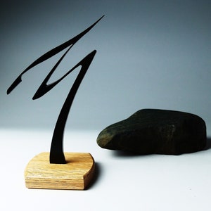 Könnte beinhalten: Eine schwarze Metallsculptur eines stilisierten Baumes auf einem Holzsockel. Die Skulptur steht neben einem dunkelgrauen Stein.