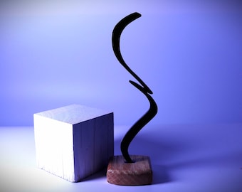 Op de afbeelding: Een zwart abstract beeldhouwwerk met een gebogen ontwerp staat op een houten basis. Het beeldhouwwerk staat op een witte kubus tegen een lichtpaarse achtergrond.