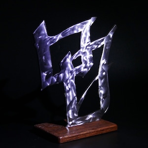 May include: A silver metal sculpture of a stylized Chinese character on a wooden base. The sculpture has a hammered texture and is illuminated by a light source.