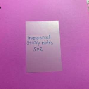Pode incluir: Um post-it transparente de 7,6 x 5,1 cm está colocado sobre um fundo roxo. O texto "Transparent Sticky notes 3 x 2" está escrito em tinta azul no post-it. O fundo parece ser um caderno.