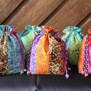May include: Six small fabric drawstring pouches featuring vertical panels of colorful batik-style floral patterns in shades of blue, brown, orange, pink, purple, and green, each finished with braided cord ties and small decorative beads at the ends.
