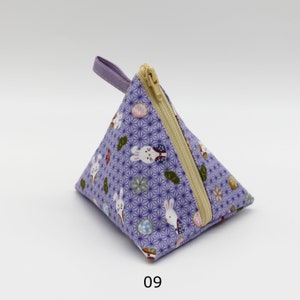 Könnte beinhalten: Ein dreieckiger, lavendelfarbener Beutel mit Reißverschluss und kleiner Schlaufe. Der Stoff weist ein sich wiederholendes Muster aus weißen Kaninchen, Blumen und geometrischen Formen auf. Der Reißverschluss ist beige, und die Schlaufe ist lavendelfarben. Die Zahl "09" ist unten aufgedruckt.