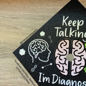 Op de afbeelding: Zwarte afstudeerhoed met glitter en witte tekst die "Keep Talking I'm Diagnosed" luidt. De hoed heeft ook een witte omtrek van een hersenen en een roze hersenen met een witte omtrek.