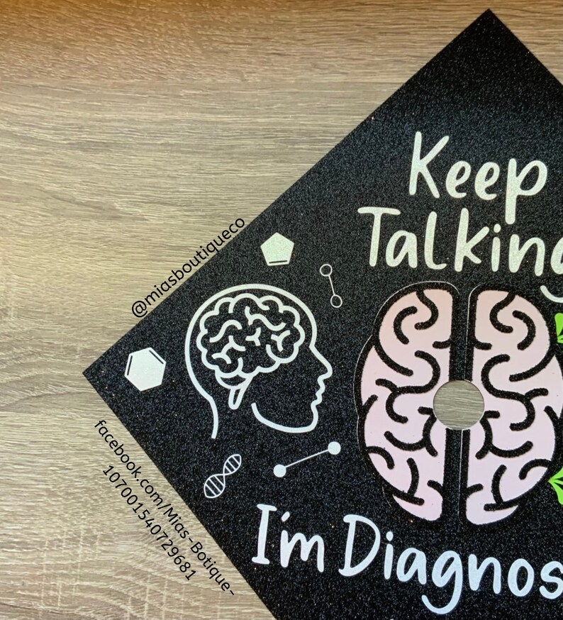 Op de afbeelding: Zwarte afstudeerhoed met glitter en witte tekst die "Keep Talking I'm Diagnosed" luidt. De hoed heeft ook een witte omtrek van een hersenen en een roze hersenen met een witte omtrek.