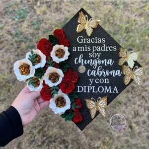 Op de afbeelding: Een zwarte afstudeerhoed versierd met rode en witte papieren bloemen, gouden vlinders en de tekst "Gracias a mis padres soy chingona cabrona y con DIPLOMA".