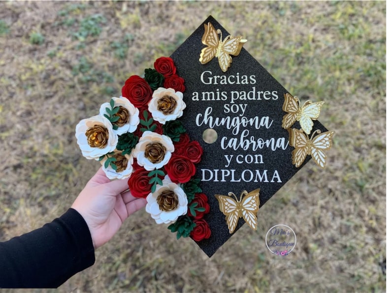 Op de afbeelding: Een zwarte afstudeerhoed versierd met rode en witte papieren bloemen, gouden vlinders en de tekst "Gracias a mis padres soy chingona cabrona y con DIPLOMA".