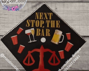 Puede incluir: Un gorro de graduación negro con el texto "Next Stop, The Bar" en letras doradas. El gorro está decorado con vasos rojos de plástico, una jarra de cerveza, copas de vino y una balanza de la justicia roja. El texto "@miasboutiqueco" y "facebook.com/Mias-Boutique 107001540729681" también está en el gorro.