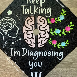 Op de afbeelding: Zwarte afstudeerhoed met glitter en witte tekst die "Keep Talking I'm Diagnosing You" luidt, met een roze en witte hersengrafiek en een groen bloemmotief. De tekst "@miasboutiqueco" staat in de rechterbovenhoek. Een wit Grieks Psi-symbool staat in de rechterbenedenhoek.