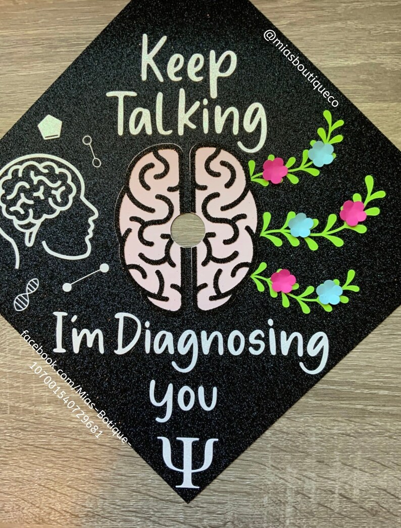 Op de afbeelding: Zwarte afstudeerhoed met glitter en witte tekst die "Keep Talking I'm Diagnosing You" luidt, met een roze en witte hersengrafiek en een groen bloemmotief. De tekst "@miasboutiqueco" staat in de rechterbovenhoek. Een wit Grieks Psi-symbool staat in de rechterbenedenhoek.