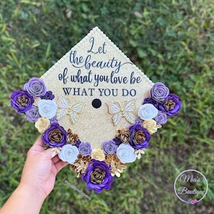 May include: A gold graduation cap decorated with purple, white and gold paper flowers. The cap has a quote that reads "Let the beauty of what you love be what you do".