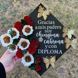 Op de afbeelding: Een zwarte afstudeerhoed versierd met rode en witte papieren bloemen en gouden glittervlinders. Op de hoed staat de tekst "Gracias a mis padres soy chingona cabrona y con DIPLOMA".