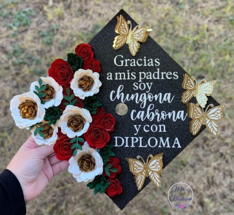 Op de afbeelding: Een zwarte afstudeerhoed versierd met rode en witte papieren bloemen en gouden glittervlinders. Op de hoed staat de tekst "Gracias a mis padres soy chingona cabrona y con DIPLOMA".