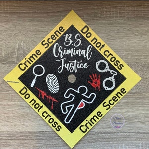 Op de afbeelding: Zwarte afstudeerhoed met gele bies en glitter. De hoed heeft een wit silhouet van een rennende persoon met een rode bloeddruppel op de borst. De tekst op de hoed luidt "Crime Scene Do not cross B.S. Criminal Justice Do not cross Crime Scene".