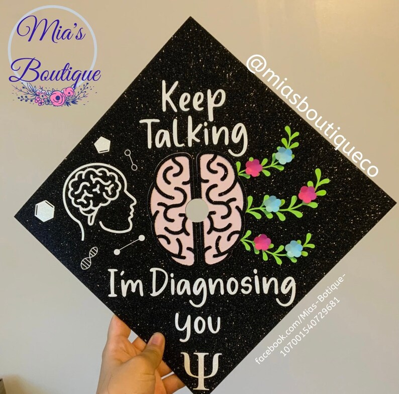 Op de afbeelding: Zwarte afstudeerhoed met glitter en een roze en witte hersenillustratie. De tekst "Keep Talking I'm Diagnosing You" is in wit geschreven. Een wit psychologisch symbool bevindt zich aan de onderkant van de hoed.