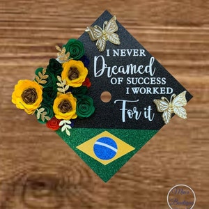 May include: Black graduation cap decorated with green and yellow paper flowers, gold glitter butterflies, and the text "I never dreamed of success I worked for it". The cap also features a green and yellow glitter background with a blue circle in the centre, resembling the Brazilian flag.