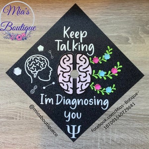 Op de afbeelding: Zwarte afstudeerhoed met een roze en wit breinontwerp. De tekst "Keep Talking I'm Diagnosing You" staat op de hoed. Een groen en wit bloemenontwerp bevindt zich aan de rechterkant van de hoed. Een witte Griekse letter Psi bevindt zich aan de onderkant van de hoed.