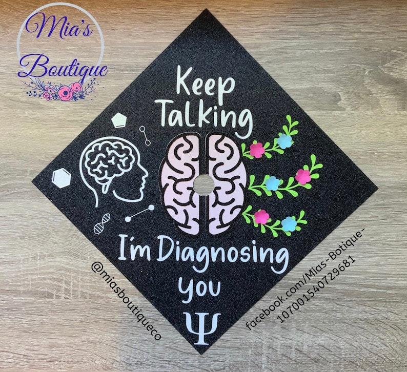 Op de afbeelding: Zwarte afstudeerhoed met een roze en wit breinontwerp. De tekst "Keep Talking I'm Diagnosing You" staat op de hoed. Een groen en wit bloemenontwerp bevindt zich aan de rechterkant van de hoed. Een witte Griekse letter Psi bevindt zich aan de onderkant van de hoed.
