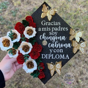 Op de afbeelding: Een zwarte afstudeerhoed versierd met rode en witte papieren bloemen, gouden vlinders en de tekst "Gracias a mis padres soy chingona cabrona y con DIPLOMA".