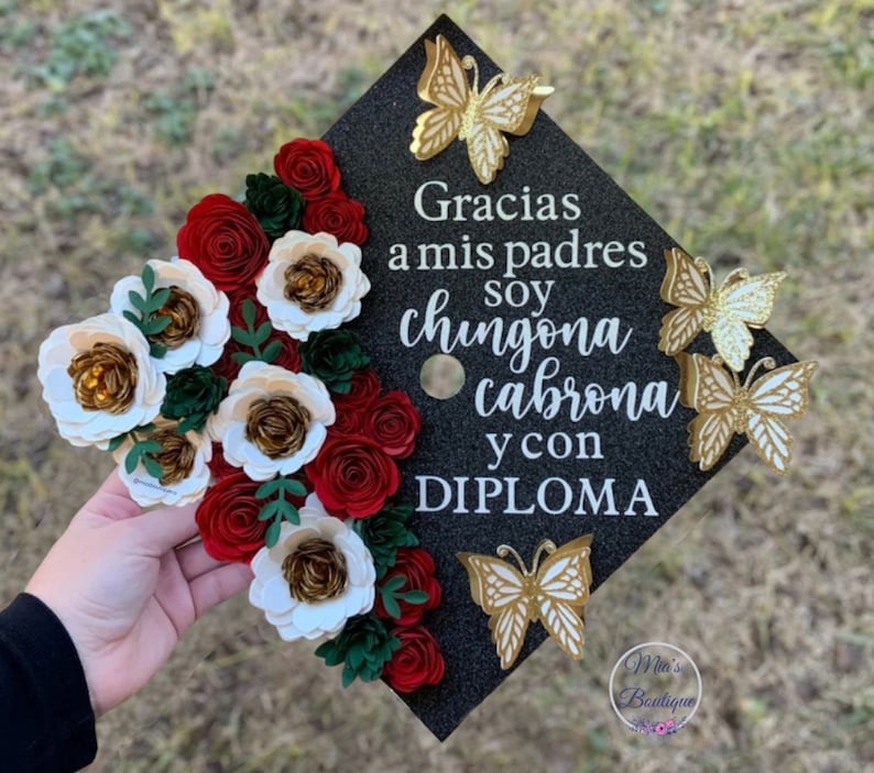 Op de afbeelding: Een zwarte afstudeerhoed versierd met rode en witte papieren bloemen, gouden vlinders en de tekst "Gracias a mis padres soy chingona cabrona y con DIPLOMA".