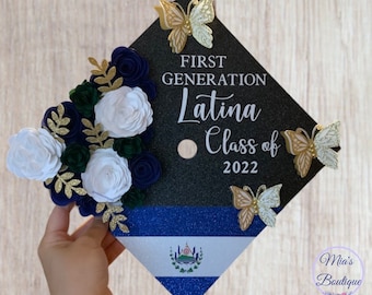 May include: A black graduation cap decorated with white and blue flowers, gold glitter, and gold butterflies. The cap also features a blue and white striped band with the flag of El Salvador. The text "First Generation Latina Class of 2022" is written on the cap.