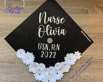 Può includere: Un cappello di laurea nero decorato con fiori di carta bianca e il testo "Nurse Olivia BSN, RN 2022".