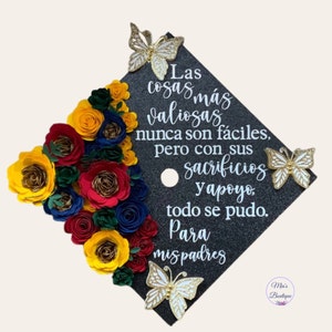 May include: A black graduation cap decorated with paper flowers in red, yellow, blue, and green. The cap has a white inscription that reads "Las cosas más valiosas nunca son fáciles, pero con sus sacrificios y apoyo, todo se pudo. Para mis padres". There are three gold and white butterfly embellishments on the cap.