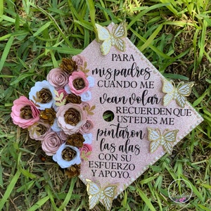 May include: A pink graduation cap decorated with paper flowers and butterflies. The cap has a message in Spanish that reads: "PARA mis padres CUANDO ME vean volar RECUERDEN QUE USTEDES ME pintaron las alas CON SU ESFUERZO Y APOYO."