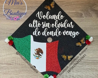May include: Black graduation cap decorated with a Mexican flag, butterflies, and flowers. The cap has the text "Volando alto sin olvidar de donde vengo" written in white.