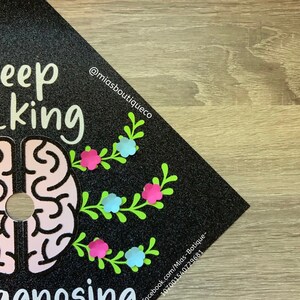 Op de afbeelding: Zwarte afstudeerhoed met glitter en een roze hersengrafiek. De tekst "Keep thinking" en "Diagnosing" staat op de hoed. De tekst "@miasboutiqueco" en "facebook.com/Mias-Boutique-1070001540729681" staat ook op de hoed.