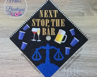 Puede incluir: Un gorro de graduación negro con purpurina dorada y pedrería. El gorro tiene un diseño de balanza de la justicia azul y el texto "NEXT STOP, THE BAR" en letras doradas. También hay imágenes de copas de vino tinto, una jarra de cerveza y vasos de plástico azules.