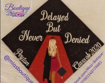 May include: Black graduation cap with gold glitter and a red and black glitter design. The cap features a silhouette of a woman walking on a red carpet with the text "Delayed But Never Denied Class of 2020".