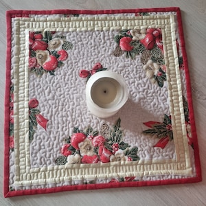 May include: A square quilted table runner with a cream background and a red border. The runner features a repeating pattern of red berries, green leaves, and white flowers. A white candle is placed in the center of the runner.